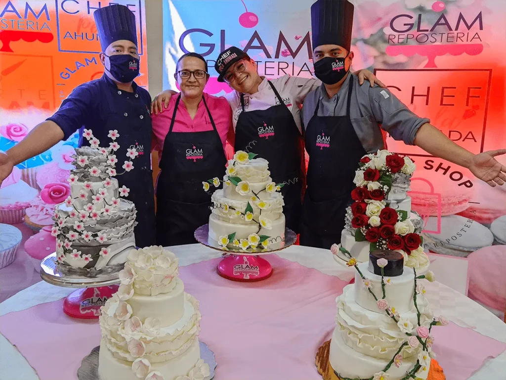Cuatro reposteros sonrientes presentan sus elaborados pasteles de boda decorados con flores en el curso de repostería avanzada.