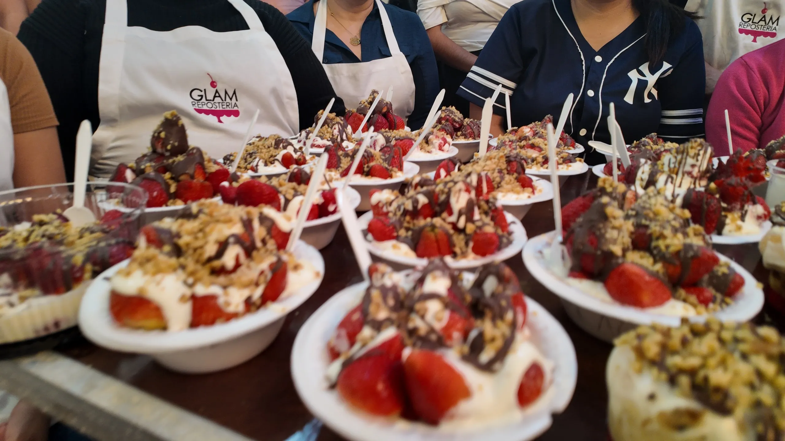 Una mesa llena de cuencos blancos con fresas, crema, chocolate derretido y nueces, con participantes del curso de fondo.