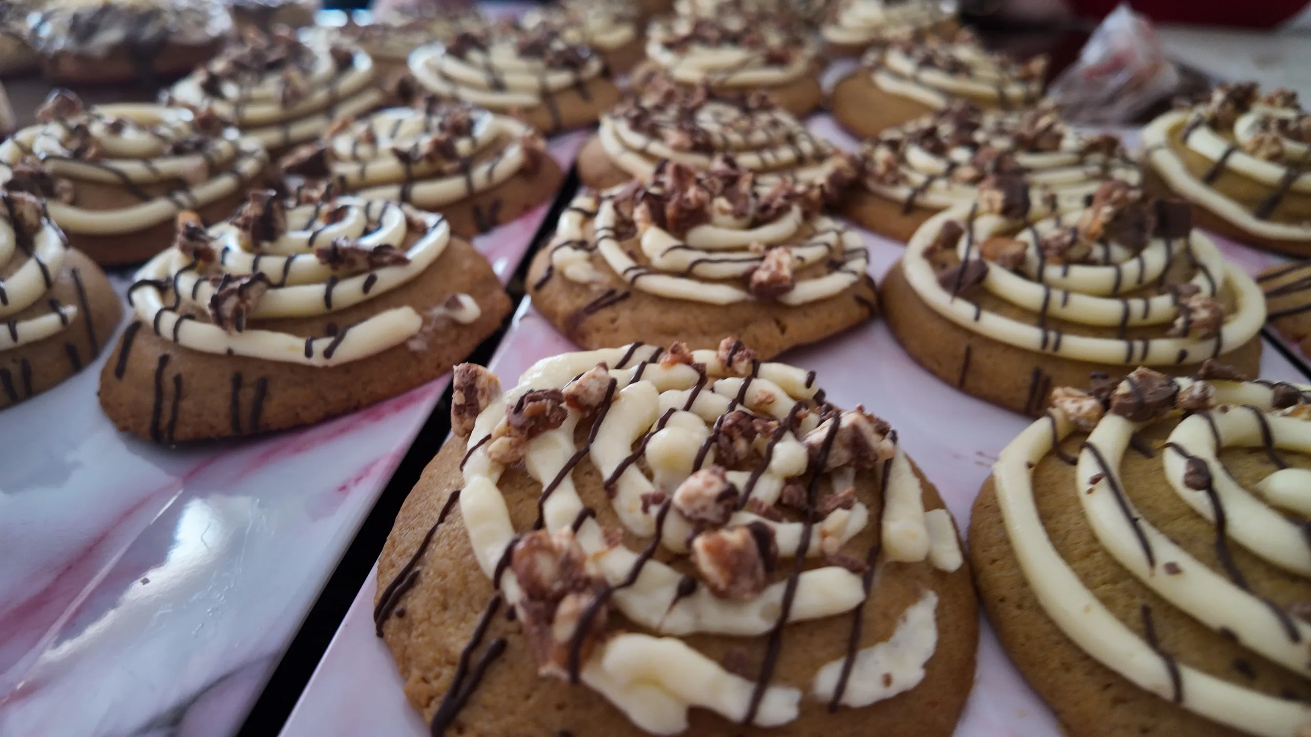 Una bandeja de mármol rosa con galletas de canela horneadas, decoradas con espirales de glaseado blanco, chocolate y nueces.
