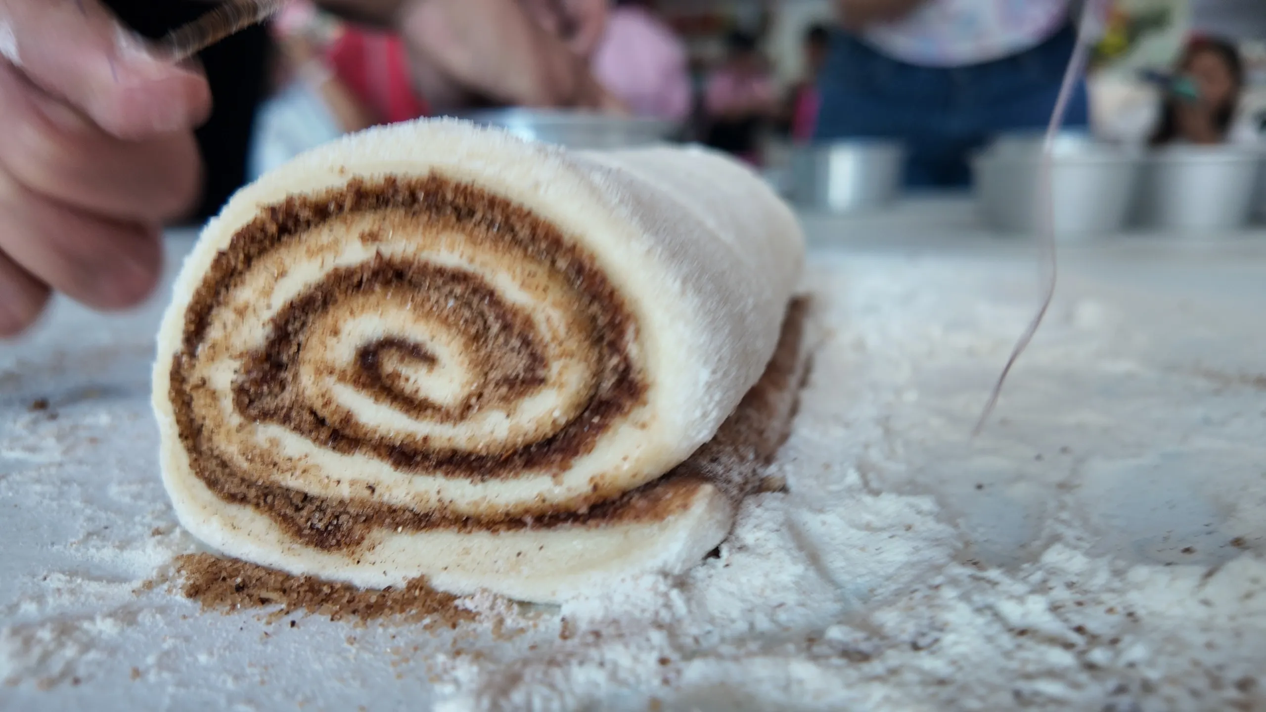 Un primer plano de un rollo de canela crudo y las manos de un estudiante aprendiendo a cortarlo con un hilo en un curso presencial.