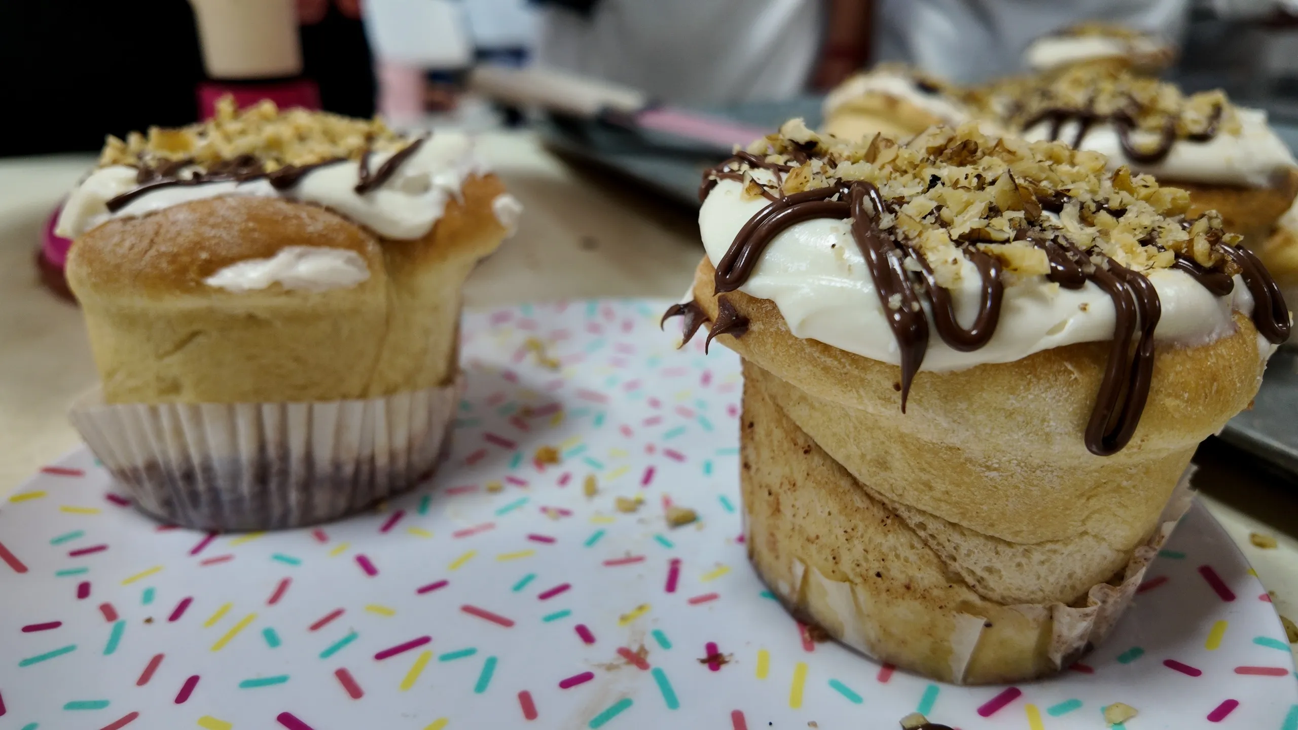 Dos muffins de rollos de canela en moldes de papel sobre un plato de lunares, decorados con glaseado blanco y chocolate.
