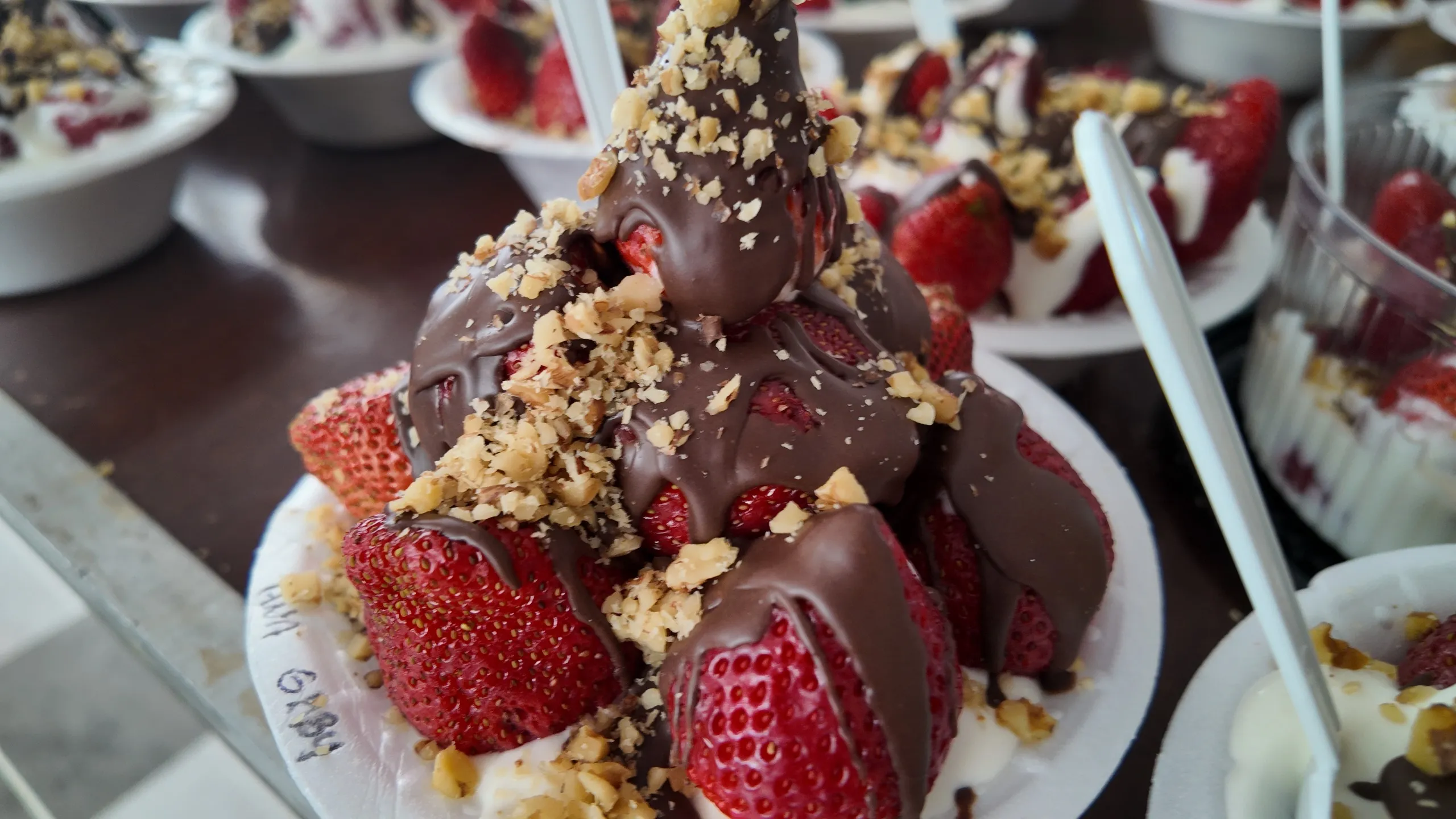Un primer plano de un postre individual en un cuenco blanco, con fresas, chocolate derretido, crema y nueces picadas.