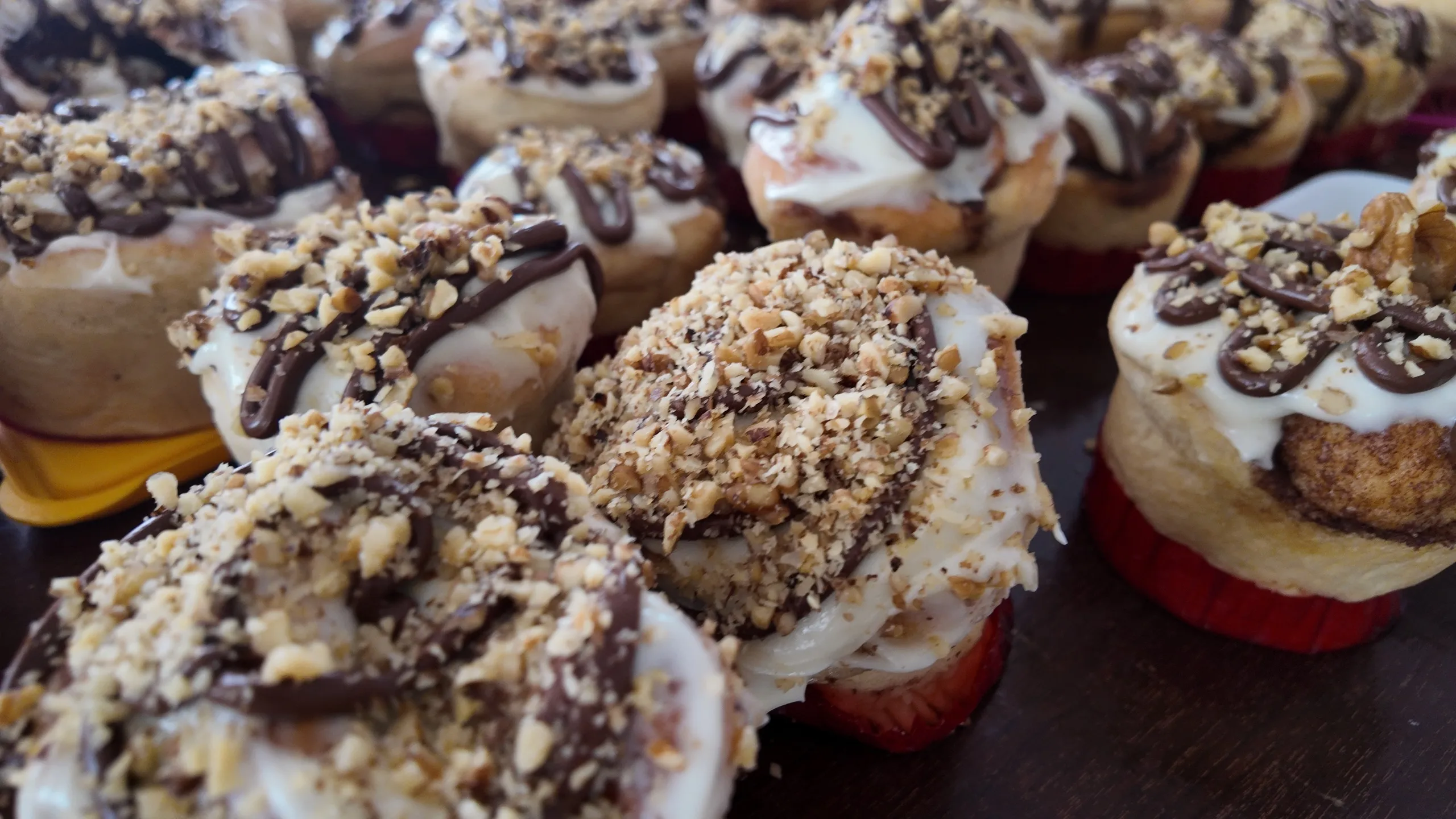 Una toma a nivel de los ojos de muffins de rollos de canela en una bandeja de metal, con glaseado, chocolate y nueces esparcidas.