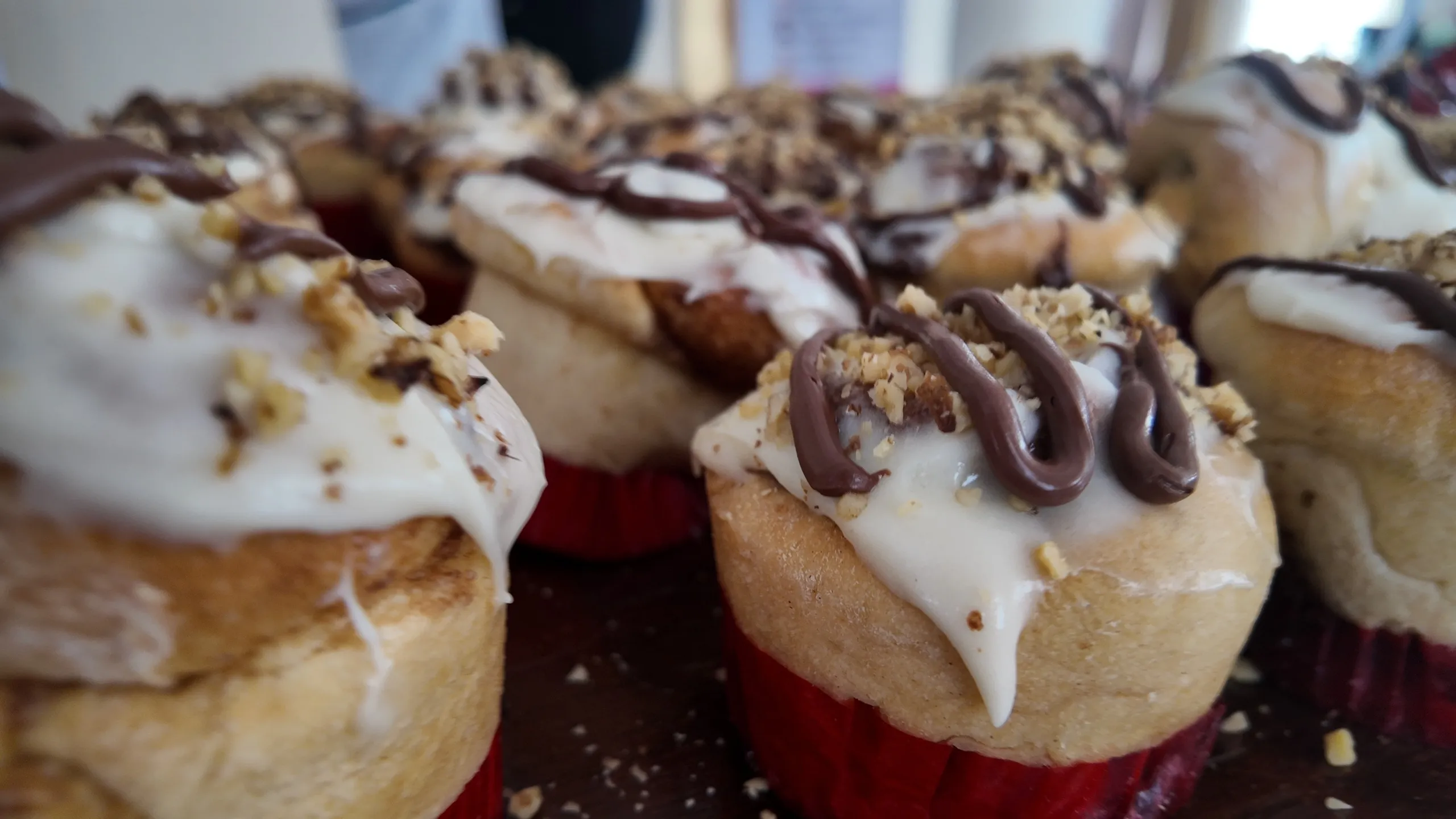 Varios rollos de canela horneados con un glaseado blanco cremoso, líneas de chocolate y nueces picadas, listos para comer.