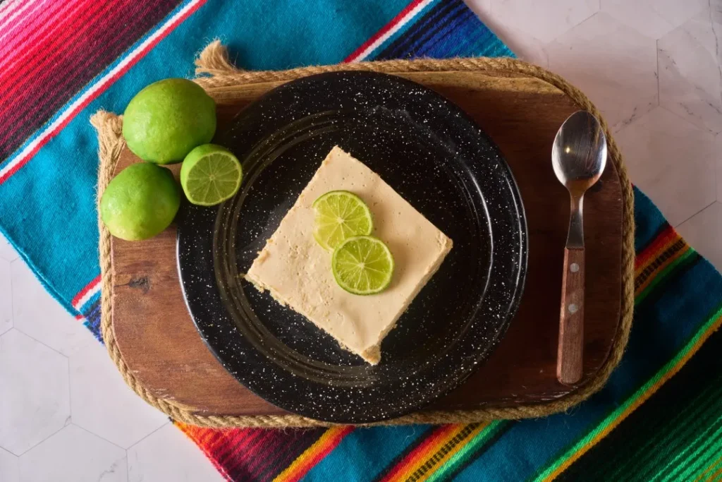 Una porción de carlota de limón en plato negro, decorada con rodajas de cítricos sobre un colorido mantel.