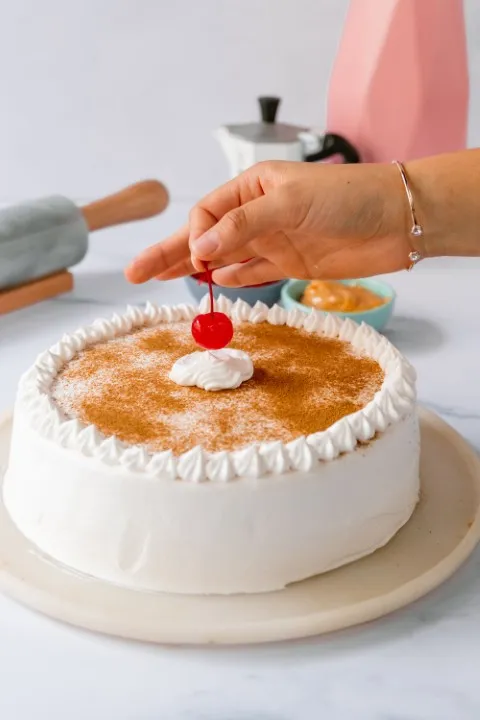 Mano colocando una cereza roja sobre un pastel beso de ángel cubierto de crema blanca y espolvoreado con canela.